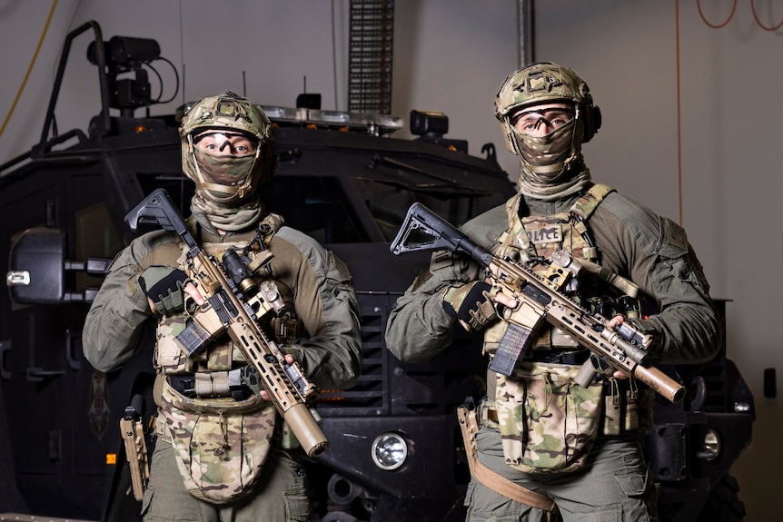 Two men in camouflaged armour hold automatic rifles and stand in front of an armoured vehicle.
