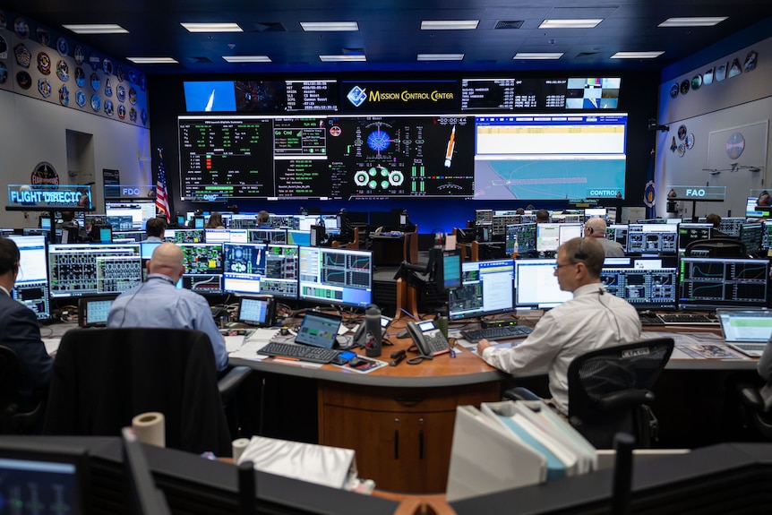 Wide photo of a control room seen from the back, with several rows of desks and screens and a wall of monitors at the front.