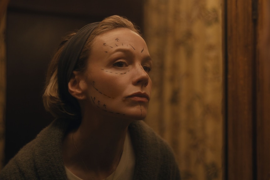 A middle-aged woman inspects her face, covered in black pre-surgery marks, in a mirror.