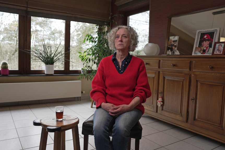 An older woman in a red jumper sits in a room, looking sad.