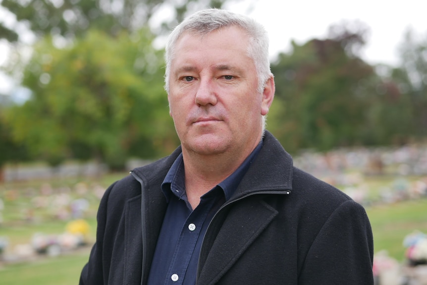 Man in a black coat and navy shirt standing in a park. 