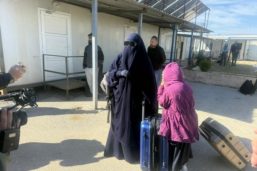 A woman wearing a black niqab and sunglasses and a small child wearing a pink parka wheeling suitcases.