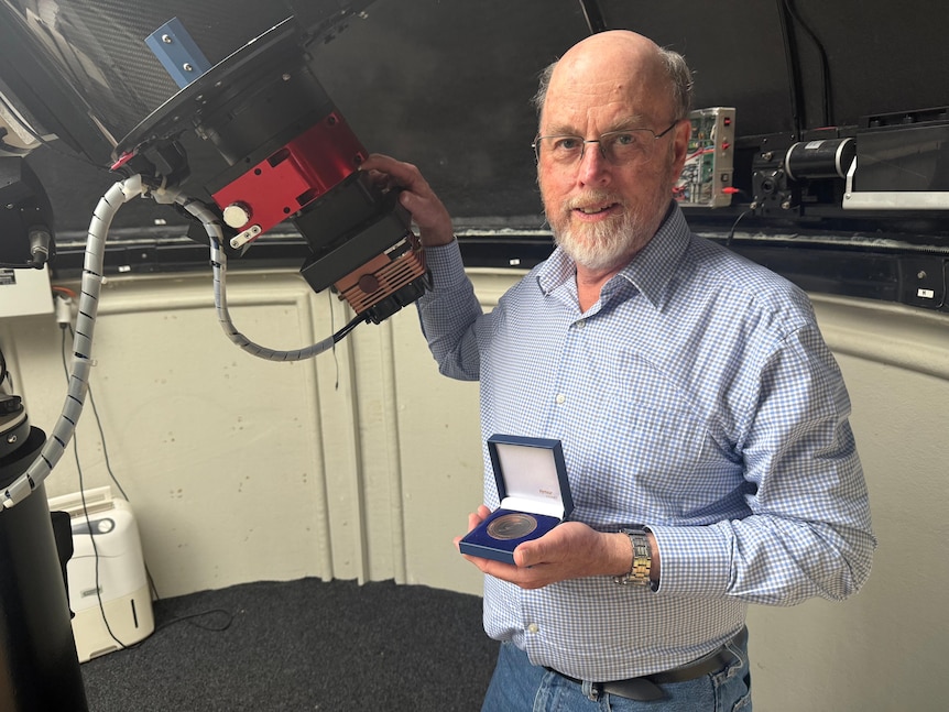A man wearing glasses and a blue button-down shirt, holding a box and grabbing a large telescope