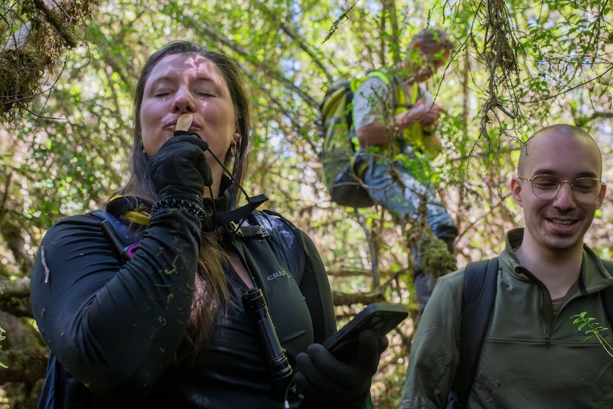 A woman with brunette hair kisses a necklace made with twine and a shell. She stands in forest. A man smiles behind her 