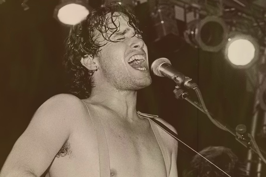 Shirtless, sweaty caucasian man passionately sings, eyes closed into a microphone on concert stage