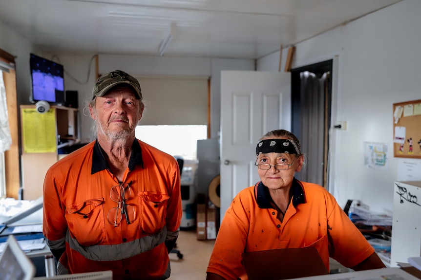 A man and woman in hi vis orange work outfits stand next to each other in an office looking straight ahead.