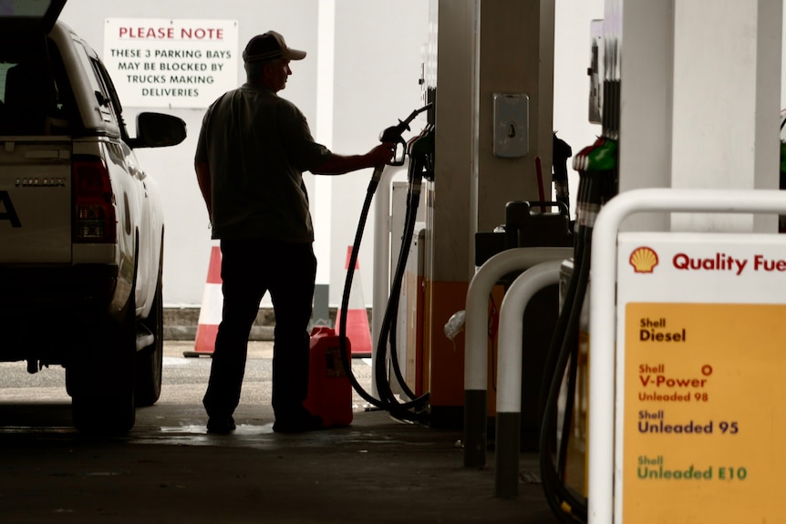 The silhouette of a man wearing a trucker cap standing next to a petrol bowser with a pump in hand.