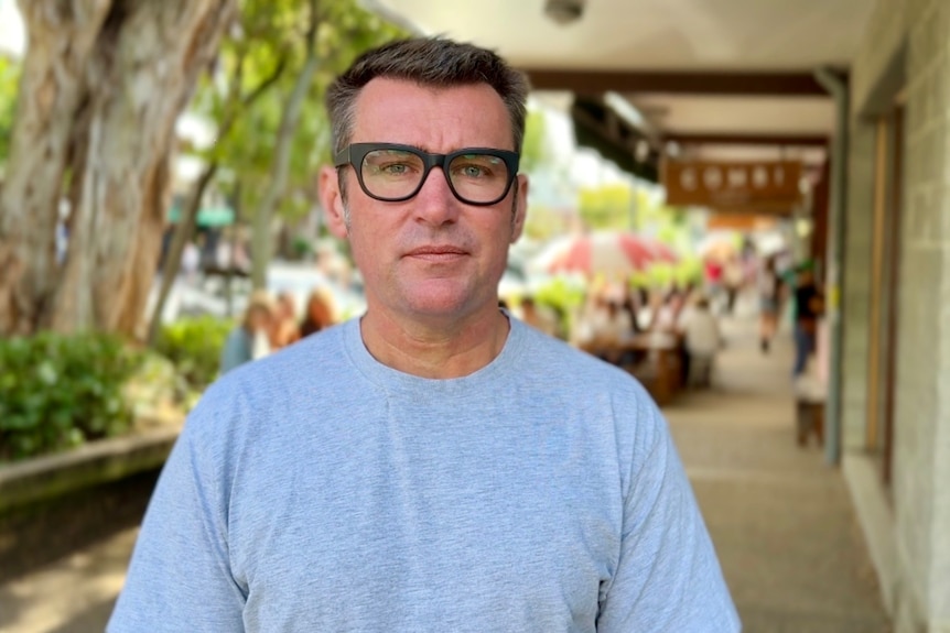 A man with short, dark hair wears glasses and a T-shirt as he stands on a footpath near a cafe.