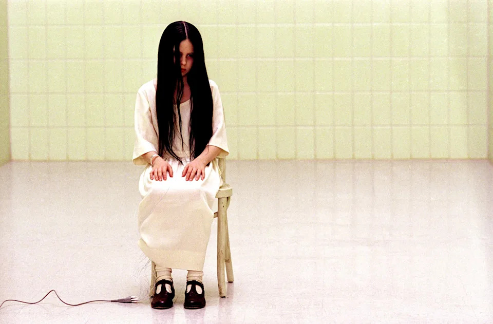 Person with long hair sits on a chair in a tiled room. There is an unplugged cable on the floor nearby