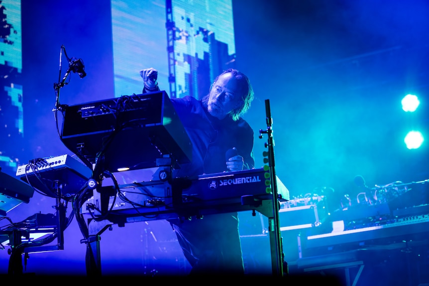 Thom Yorke reaches for a knob on a raised piece of equipment, performing live on stage, bathed in blue light