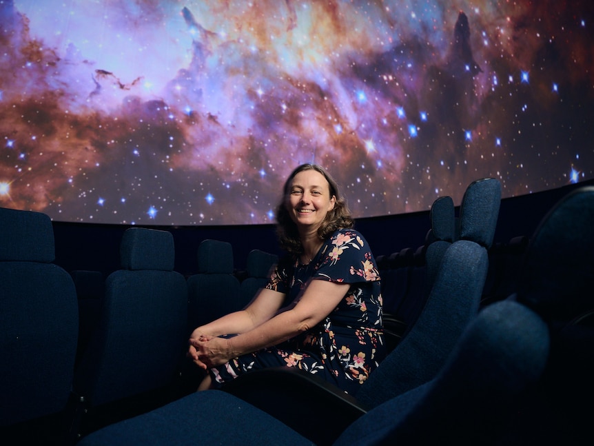 A woman wearing a floral top, sitting on a cinema chair in a cinema room, with a galaxy backdrop behind her