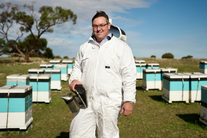 A man standing between boxes of beehives in a beekeeping suit, smiles to camera.