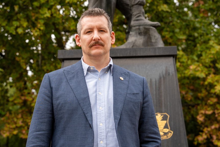 A man with a moustache standing in front of a war memorial