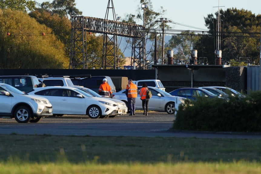 Workers in high-visibility clothing walking near parked cars in an industrial area with power poles and trees in the background.
