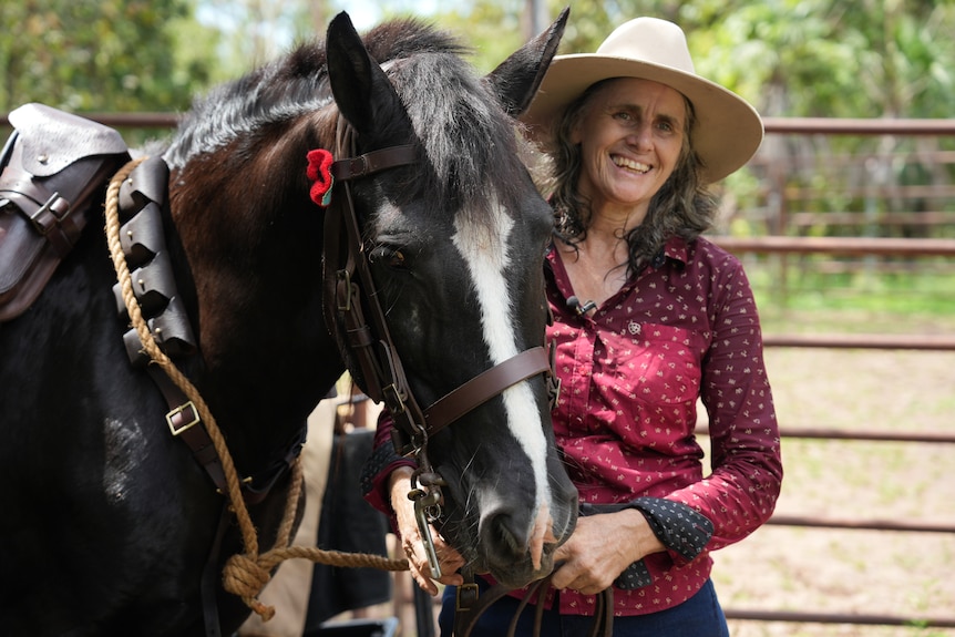 A woman, smiling widely, standing next to a horse with a red poppy tucked behind its ear, in a horse pen.