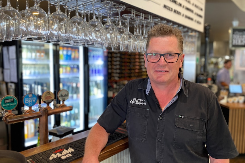 A man wearing glasses and a black collared shirt stands next to a pub bar.