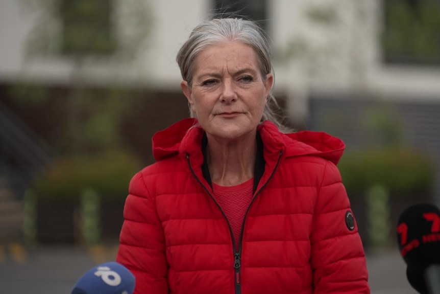 Woman wearing red puffer jacket standing in front of microphones.