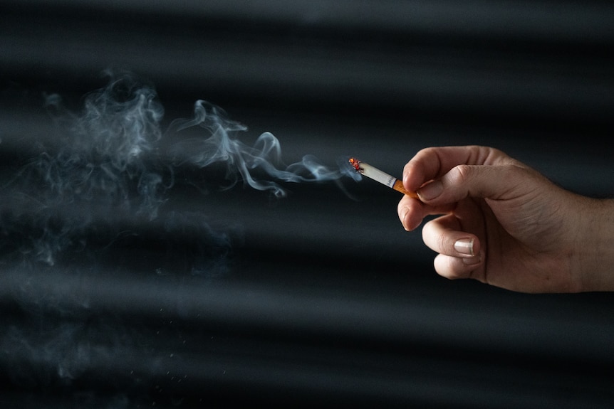 A male hand holding a smoking cigarette, in front of dark background.