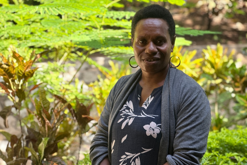 A woman smiles standing in front of green palm trees wearing a grey and black shirt