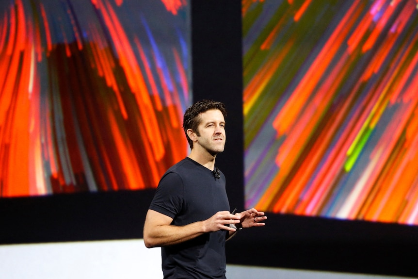 A man wearing a black top speaking on stage with a microphone