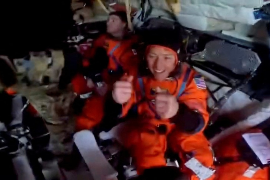 Two astronauts in orange space suits are seen sitting in a space capsule. One of them is giving a thumbs up sign.