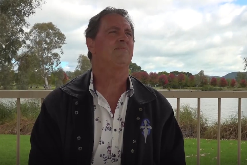 A middle-aged man in a black jacket stitting by a river.