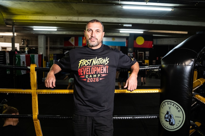 A man with short dark hair leans on the ropes of a boxing ring.