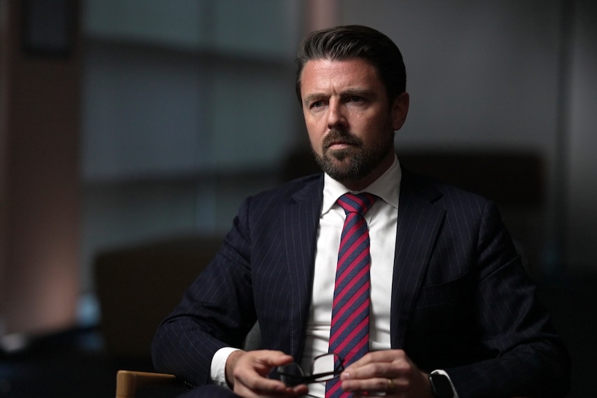 A man wearing a suit and tie sitting in a slightly darkened room. His expression is serious.