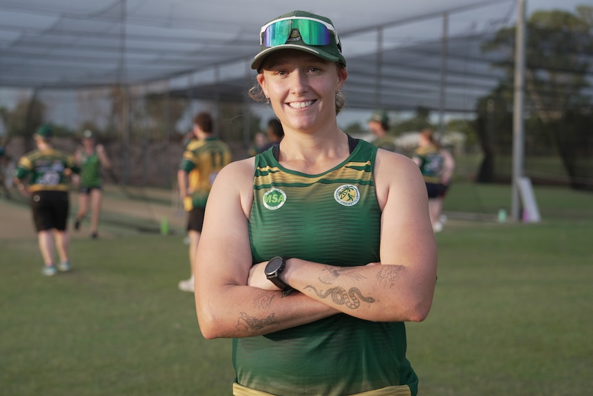 A woman wearing a green and gold sports singlet smiles at the camera, a pair of sunglasses are sitting on the cap she's wearing.