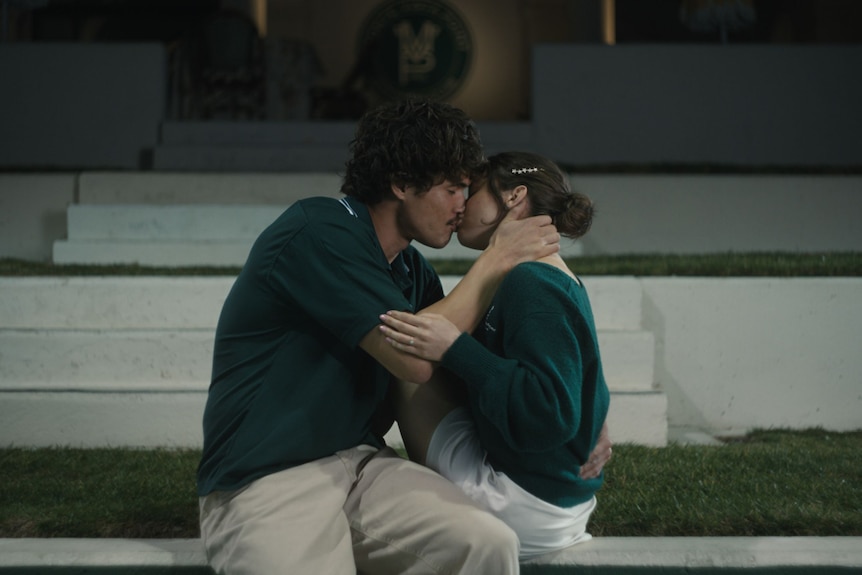 A young couple, both wearing green jumpers, kissing as they sit on a whitewashed wall in front of green lawn.