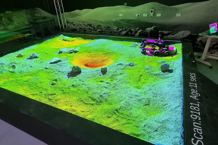 A bed of dust in green and blue light with a rover and rocks on top.