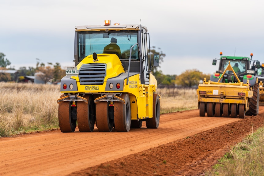 Two rollers compact dirt on a red earth road that traverses farmland.