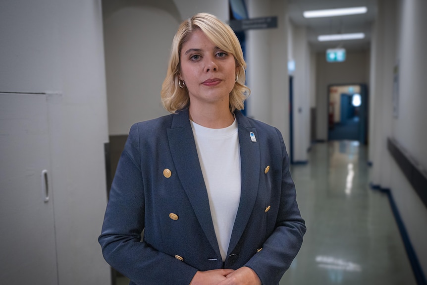 Katharine stands in a long corridor, is blonde, wears white tee, blue jacket with gold buttons.