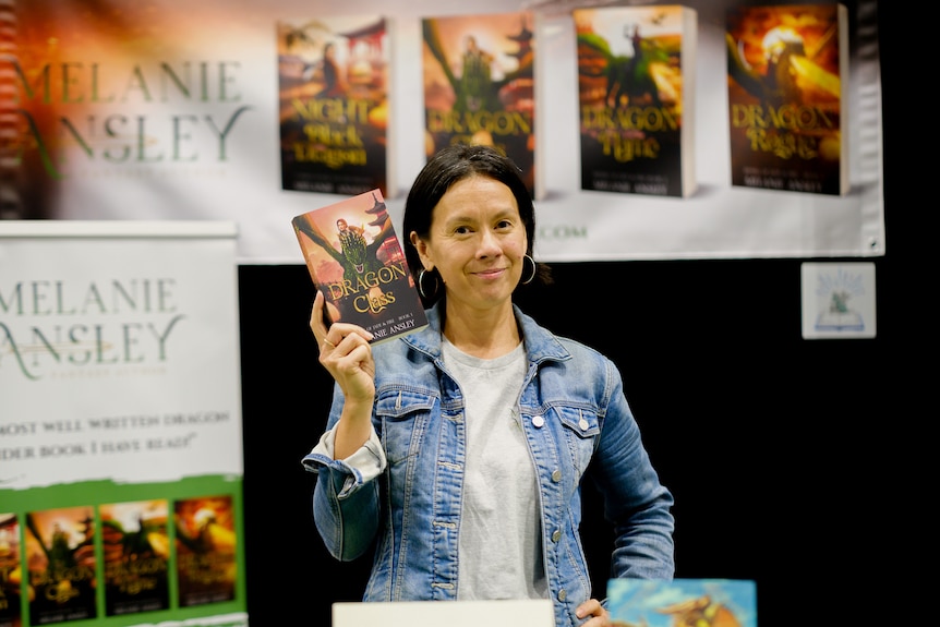 Woman holding up a book in front of a poster featuring Riders of Jade & Fire fantasy novels.