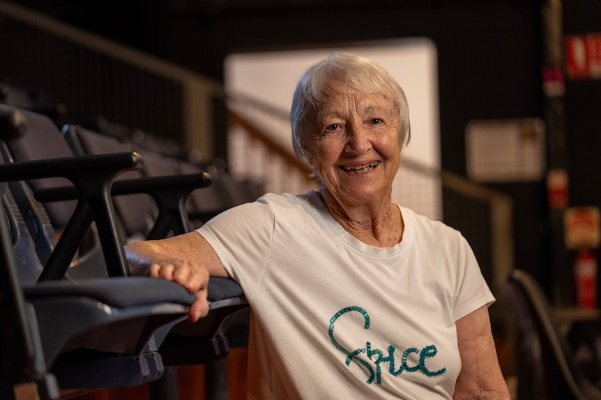 An older woman in a while shirt with short grey hair sits back in a chair in a theatre.
