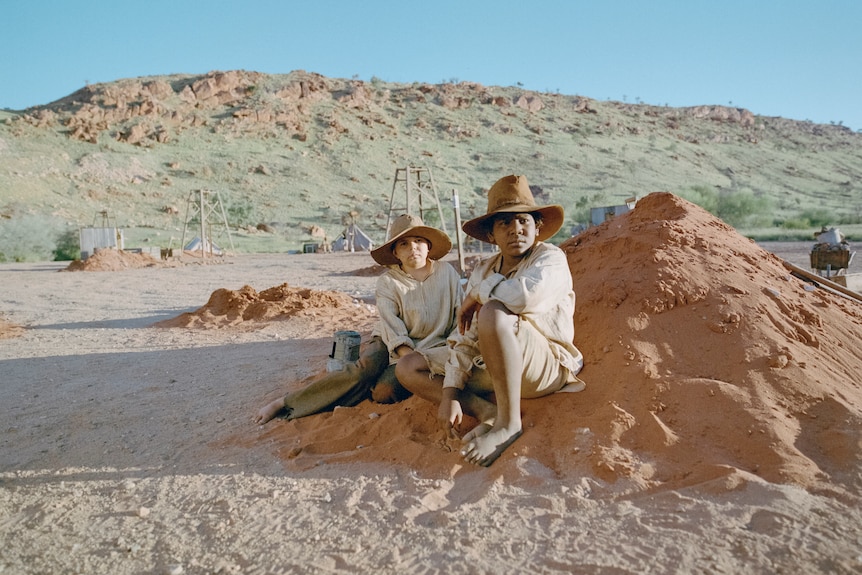 Two young boys sit on a pile of red dirt in the Australian outback