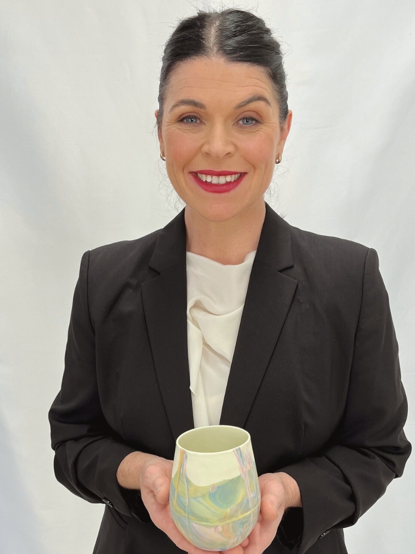 Michelle Carr, a middle-aged woman, in a blazer, one eyebrow arched, smiles, holding a ceramic cup in her hands.