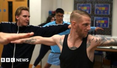 Adam Townsend is striking a yoga pose, where his arms are outstretched. He is heavily tattooed, with a brown beard. He is wearing a black vest. Other members are doing the same pose in the background.