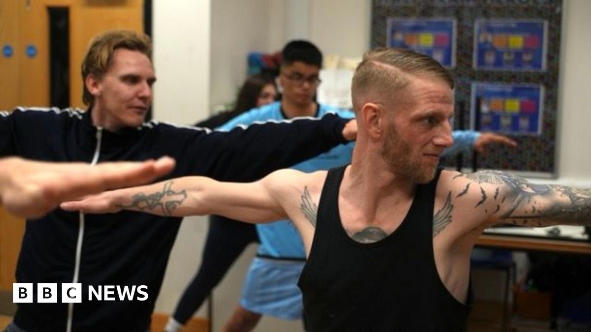 Adam Townsend is striking a yoga pose, where his arms are outstretched. He is heavily tattooed, with a brown beard. He is wearing a black vest. Other members are doing the same pose in the background.