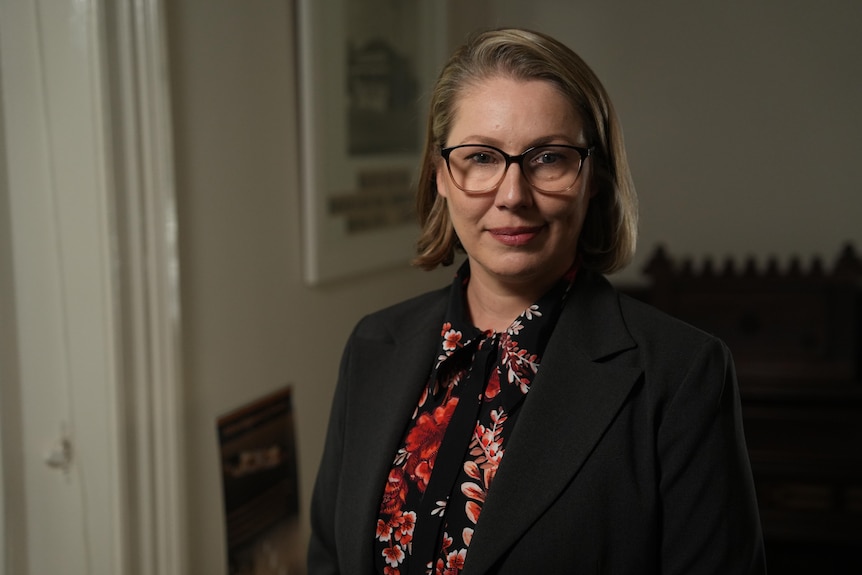 A woman with glasses in a dark room in front of a coil heater.