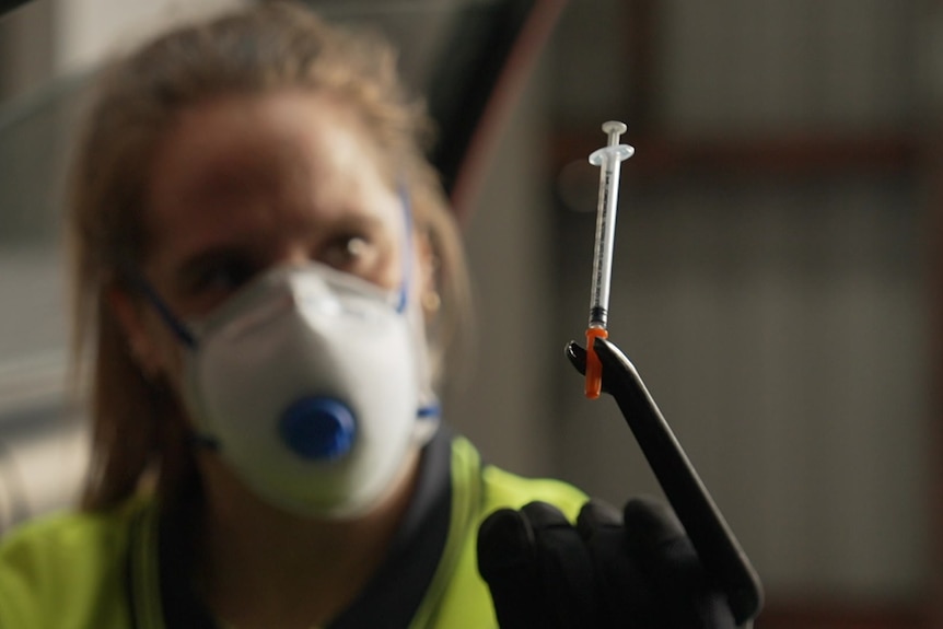 A woman wearing a face mask holds up a syringe which is in focus with an open car door in the background.