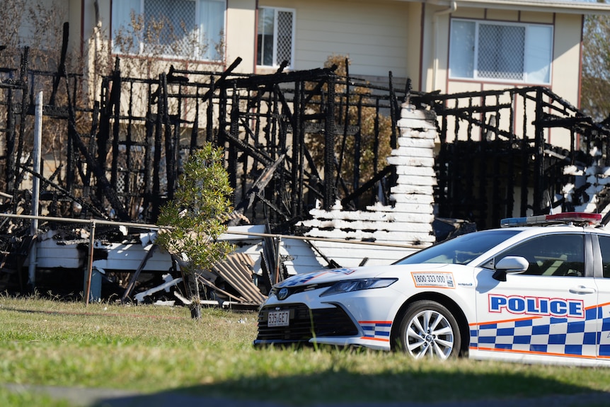 A police vehicle sits parked in front of a scorched structure. All that remains are black pieces of timber and a small wall.