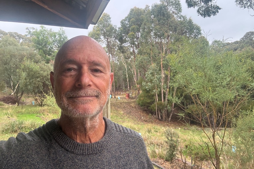 A man in a woolen jumper poses for the camera at a rural property