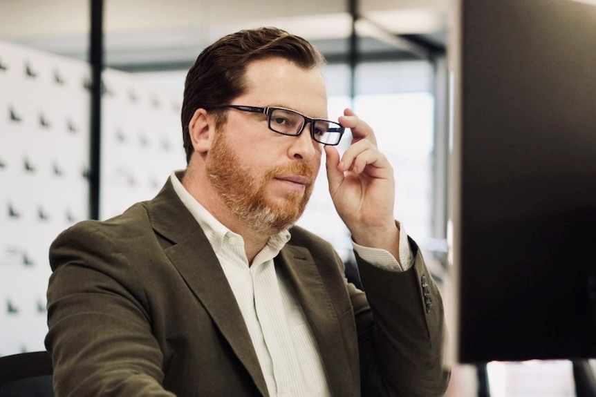 A dark-haired man looks at a computer monitor, with his hand holding the side of his glasses, which he is wearing.