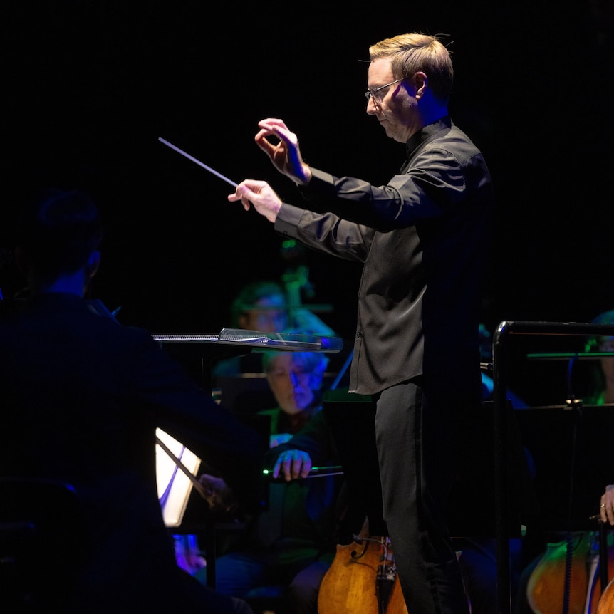 Benjamin Northey viewed from the side conducting the Sydney Symphony on a darkened stage. Both arms are held aloft.