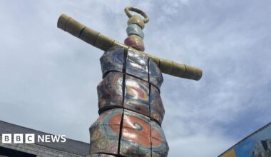 The image shows the top of a very tall statue called the Earth Goddess. It resembles a brightly coloured totem pole and has yellow arms. The sky behind it is blue with thin white cloud. The tops of two buildings are visible too.