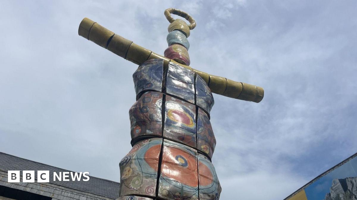 The image shows the top of a very tall statue called the Earth Goddess. It resembles a brightly coloured totem pole and has yellow arms. The sky behind it is blue with thin white cloud. The tops of two buildings are visible too.
