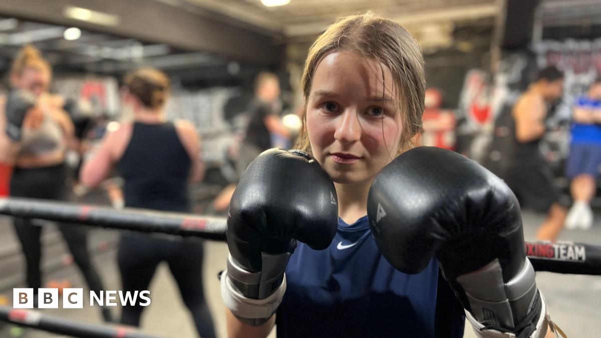 A 19 year-old woman is standing next to a boxing ring. She is wearing black boxing gloves which she is holding up close to her face. She has light brown hair tied back and is wearing a navy T-shirt. Other poeple are training in the ring behind her.