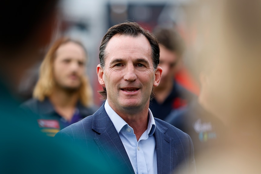 AFL executive Andrew Dillon speaks to the media, framed by blurry figures in the foreground..