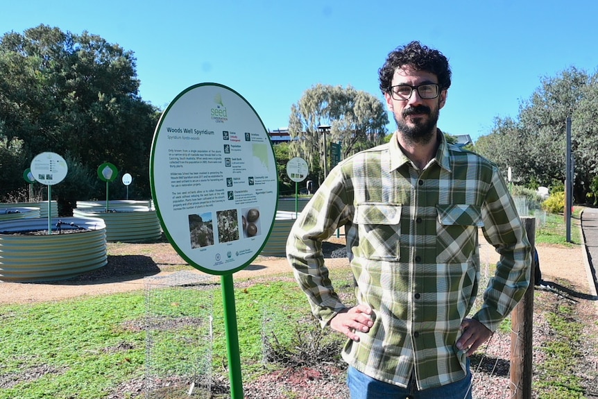 A man in a flannel shirt stands next to plants and signage